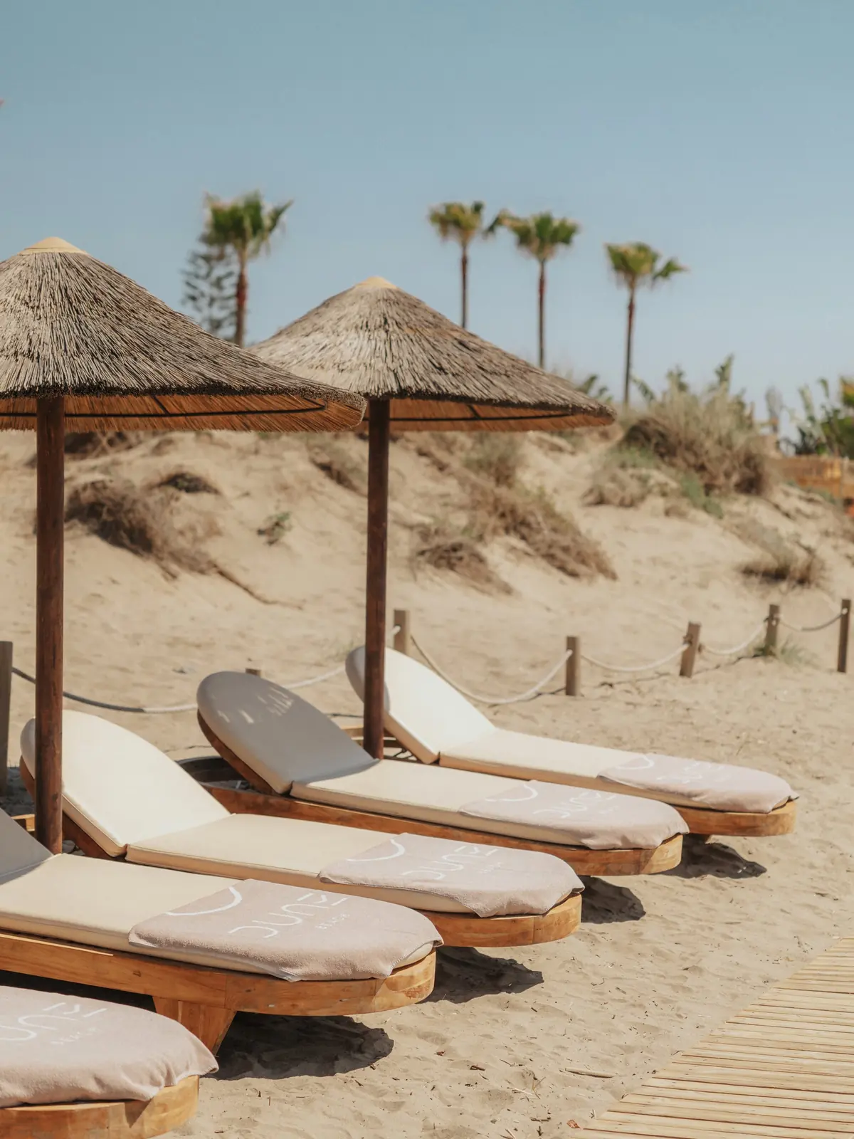 Wooden beds with Dune towels under thatched parasols, palms and dunes behind