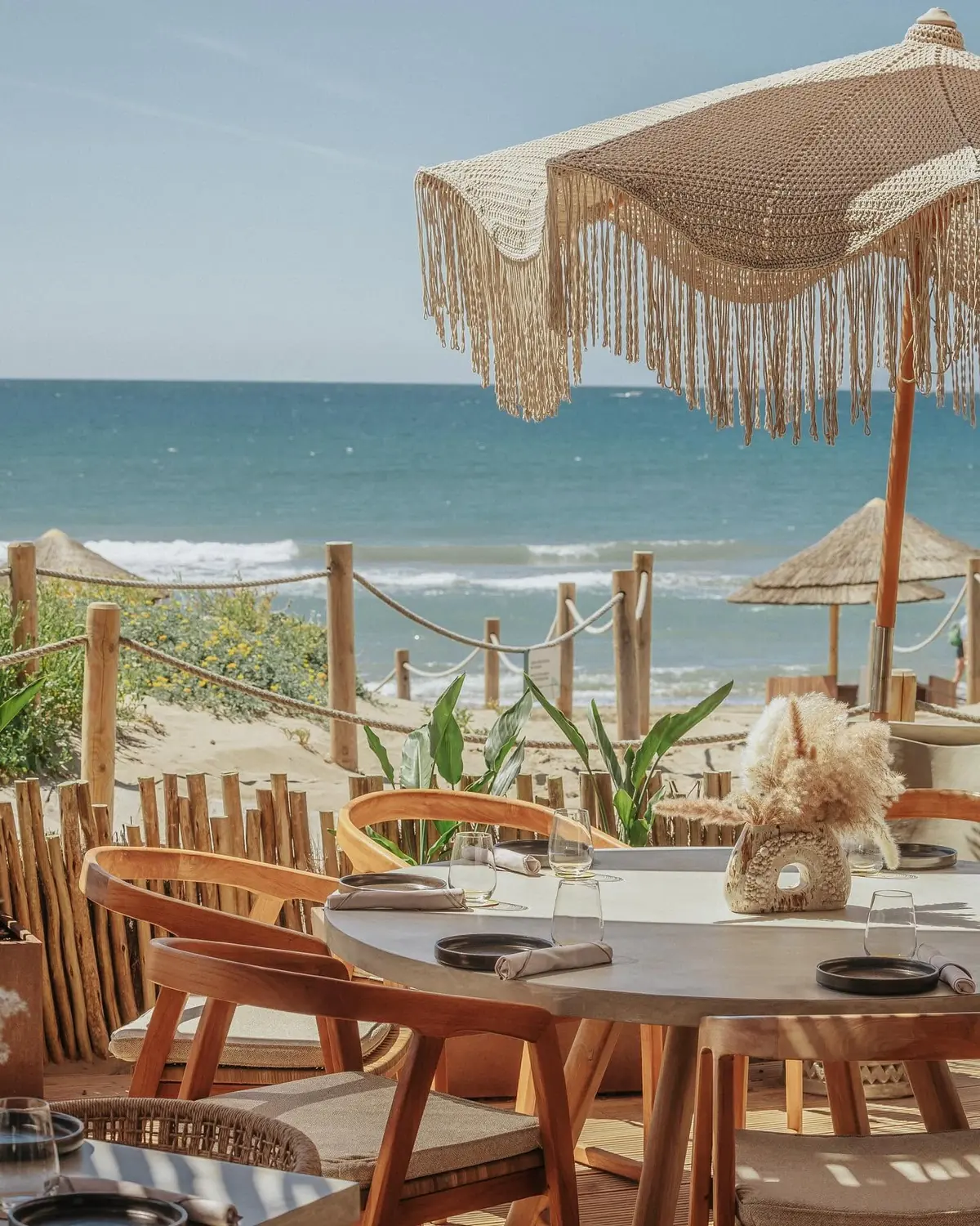 A laid table on the terrace under a macramé parasol, the sea beyond